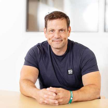 David Beard, Head of Technology for BWBF, sits at a table smiling with his hands resting on the table in front of him. He has short brown hair and is wearing a dark t-shirt.