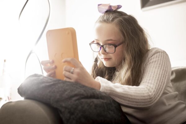 A girl is sitting on a sofa with a Voxblock audio book player in her hands. She is wearing glasses and has a bow in her hair.