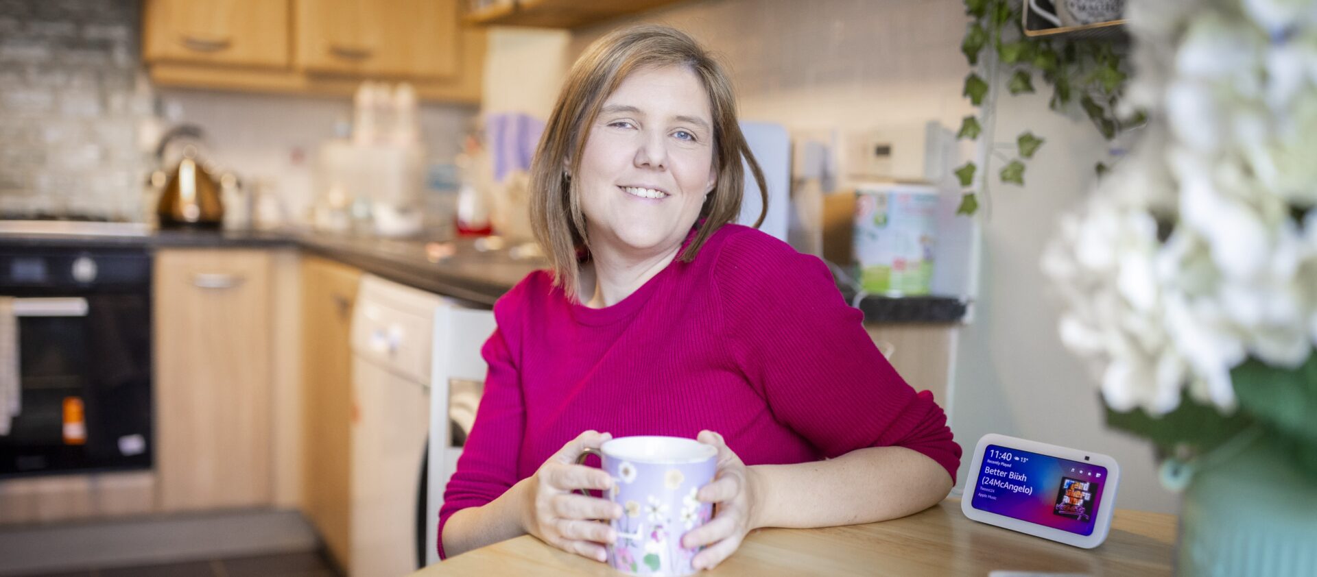 A young woman, Laura, sits at a kitchen table with a cup of tea. She is smiling and wearing a vibrant pink top. On the table next to her is an Echo Show Alexa Device.