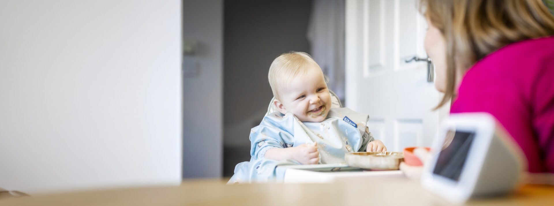 Baby Daphne is sitting in a high chair with a bowl of food in front of her. She is wearing a bib and smiling broadly. In the foreground her mother, Laura, is leaning towards her. There is an Echo Show Alexa device on the table in the foreground.