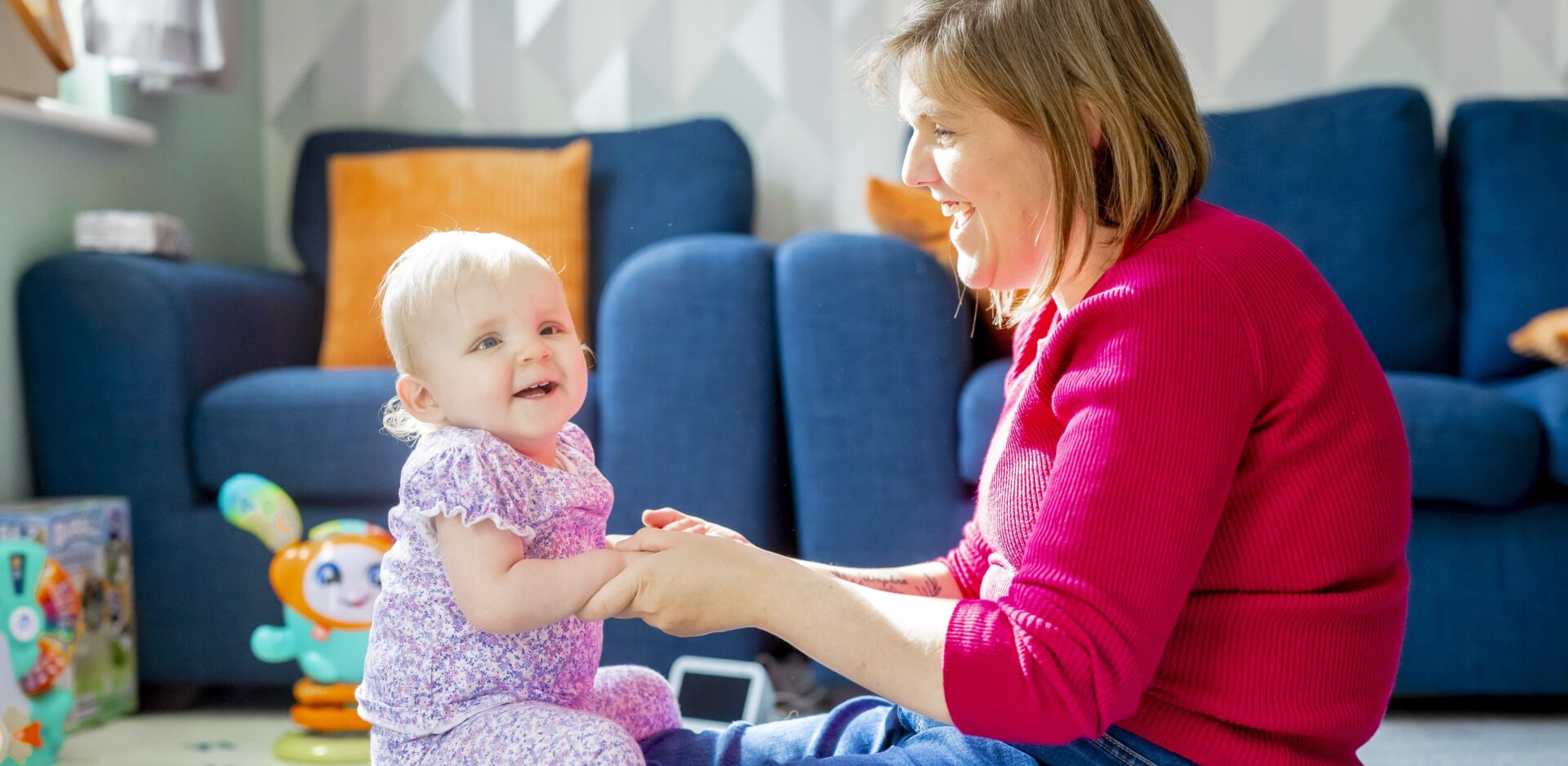 Mum, Laura, and her baby daughter, Daphne sit facing each other and holding hands in a living room filled with children's toys. Both are smiling. On the floor is an Echo Show Alexa device.