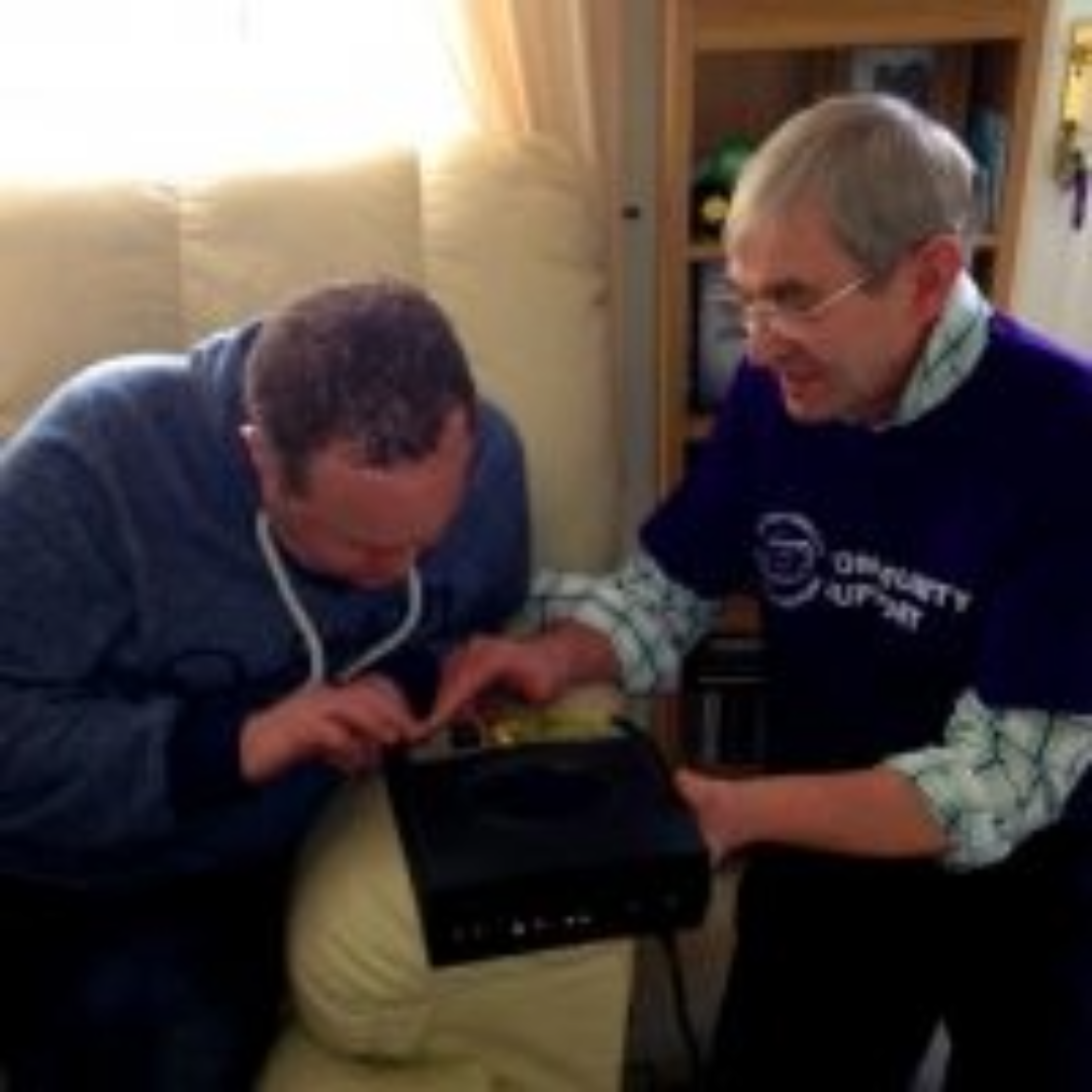 A BWBF volunteer in a purple top is helping a person to use an audio device. Both are leaning into the device interacting with the buttons.