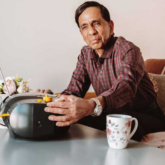 BWBF service user Krisha sits on a sofa. His hand is resting on his Concerto 3 radio from BWBF, which is on the table in front of him.