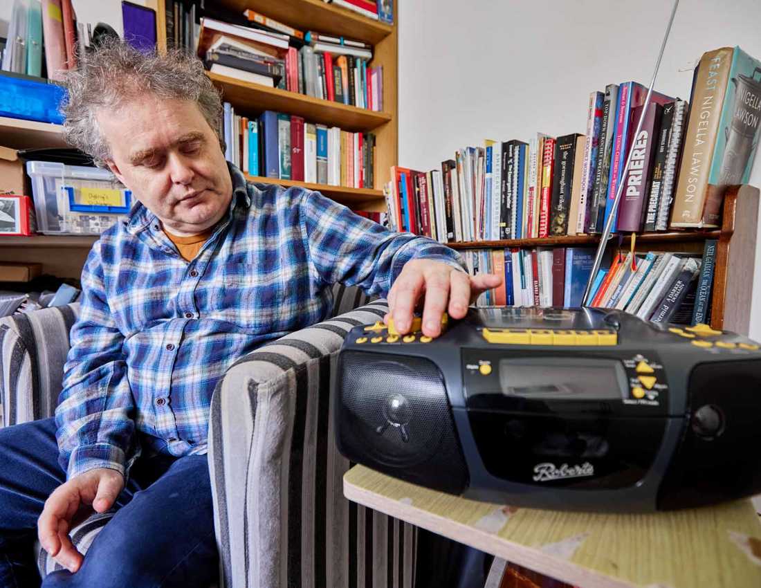 BWBF service user Dyfrig sits in a chair in a room with bookshelves. His Concerto 3 radio from BWBF is on a table next to him