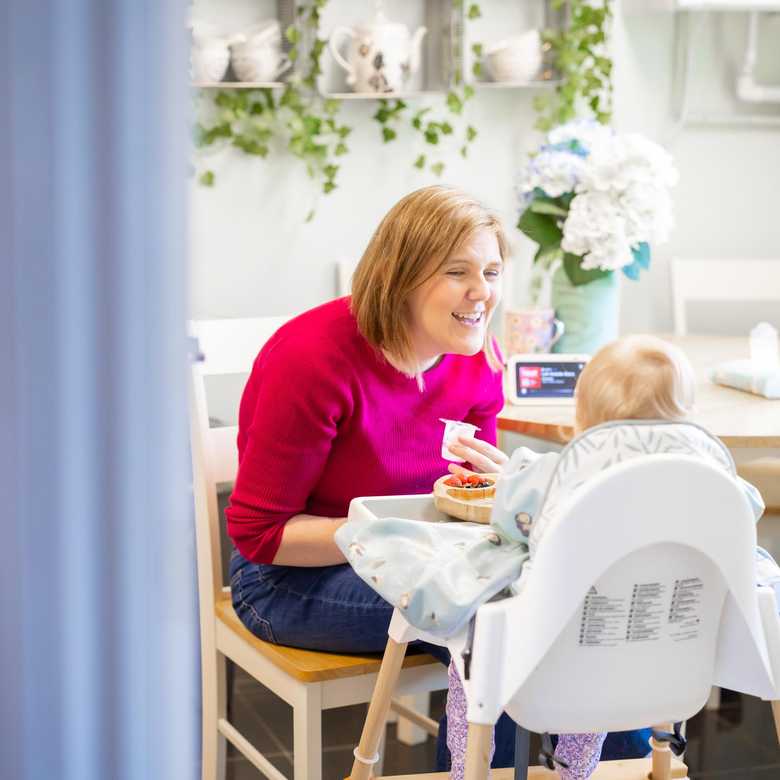 Laura and baby Daphne in their kitchen. Daphne is in a high chair and Laura is smiling as she feeds her baby. On the table is the Alexa Echo Show device BWBF gave them.