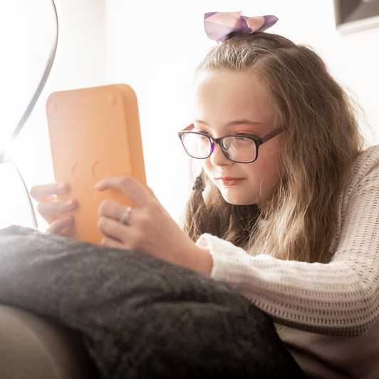 Annabel sits on her sofa reading her Voxblock audio book player. She wears glasses and has a bow in her hair.
