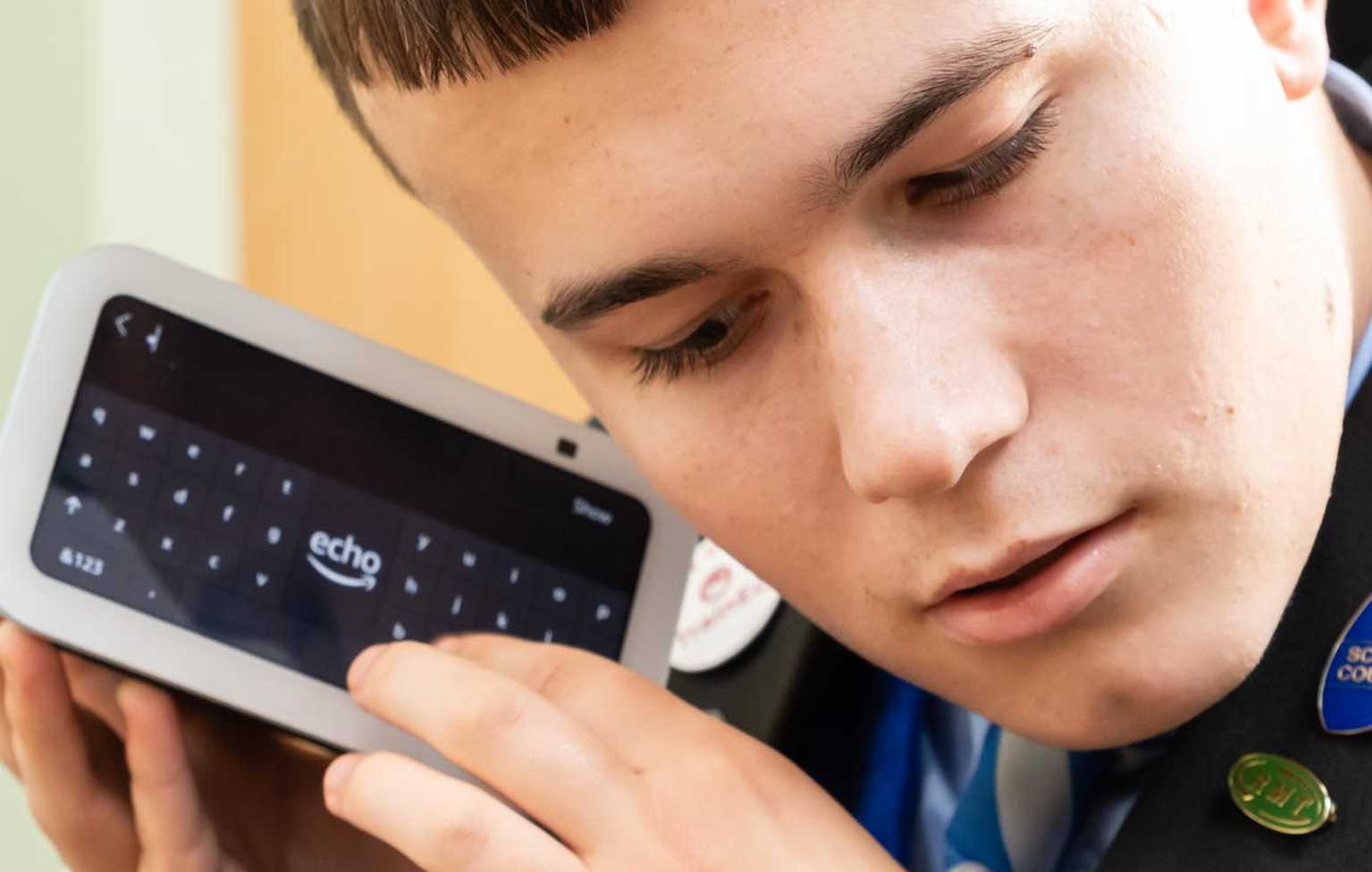 A young man wearing a school blazer holds a white Amazon Echo Show device up to his ear.
